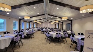 Pine Hall has an arched ceiling. Tables and chairs are set up for a wedding, with lilac chair bows and lilac centerpieces.